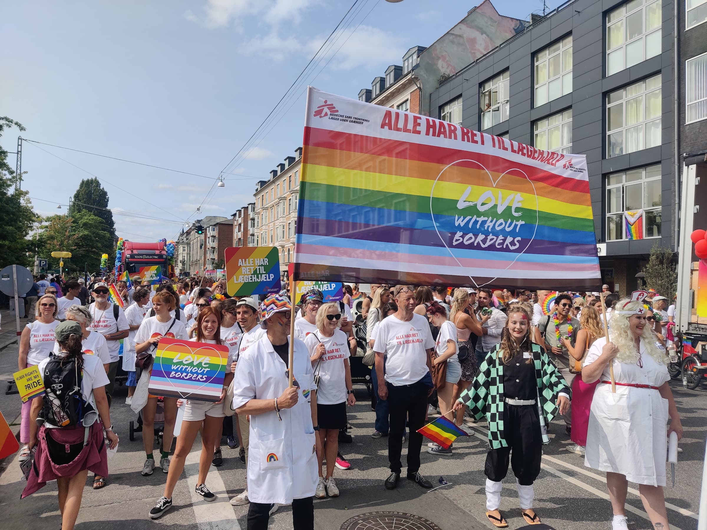 Store og små grupper fra Læger uden Grænser marcherer til Copenhagen Pride med regnbuefarvede bannere og skilte med teksten “Love Without Borders” og “Alle har ret til lægehjælp,” i støtte til LGBTQ+ rettigheder, omgivet af mennesker i farverige outfits og kostumer.